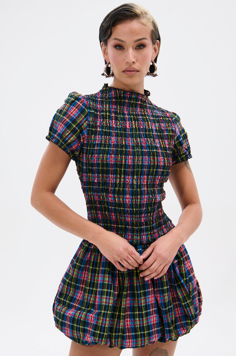 A woman models the THAT GIRL PLAID MINI DRESS—a short, fitted plaid mini with puffed sleeves and a ruffled high neckline—standing against a plain white background, her short slicked-back hair and statement earrings completing the look.