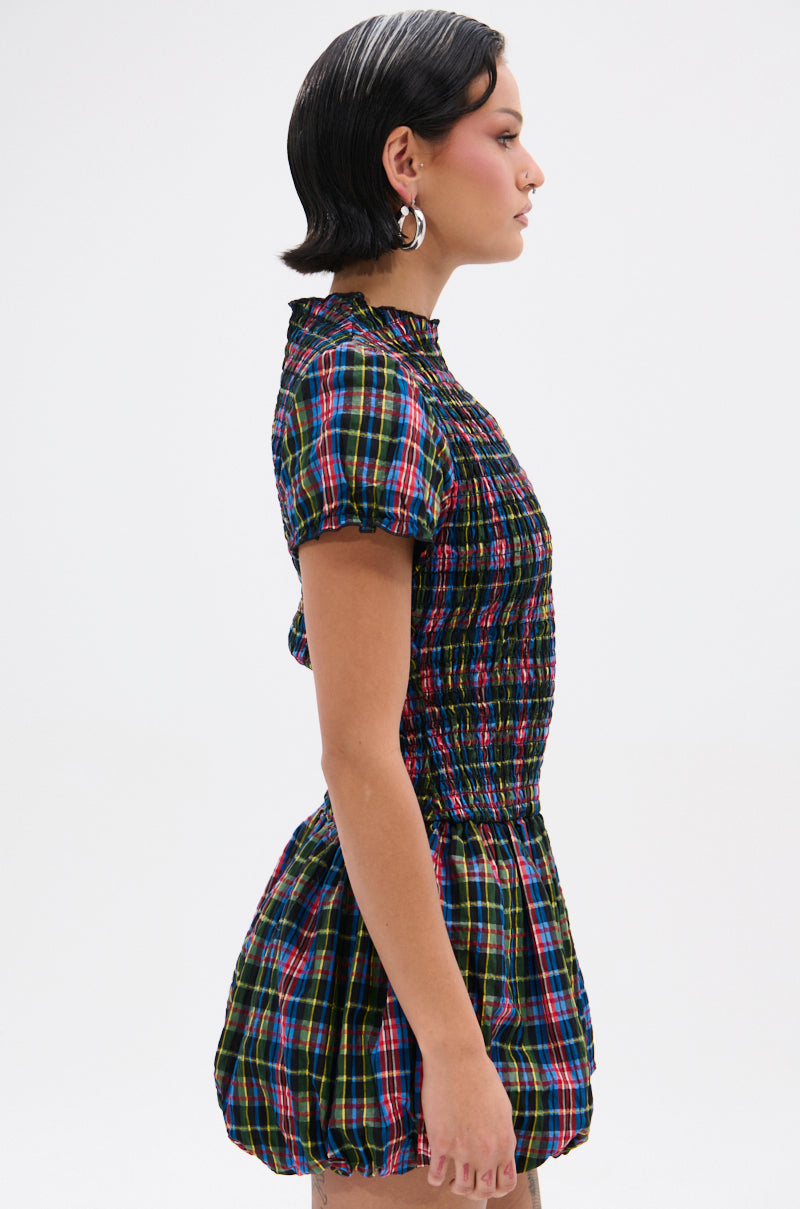 A person with short, dark hair wears hoop earrings and the THAT GIRL PLAID MINI DRESS with a smocked bodice, short sleeves, and bubble skirt, standing in profile against a plain light background.