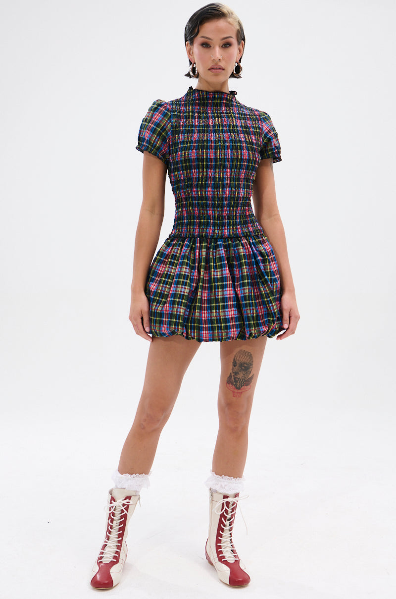 A person stands on a white background wearing the THAT GIRL PLAID MINI DRESS with a smocked bodice and short puff sleeves, paired with red and white lace-up boots. They have short hair and a visible leg tattoo.