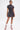 A person stands on a white background wearing the THAT GIRL PLAID MINI DRESS with a smocked bodice and short puff sleeves, paired with red and white lace-up boots. They have short hair and a visible leg tattoo.