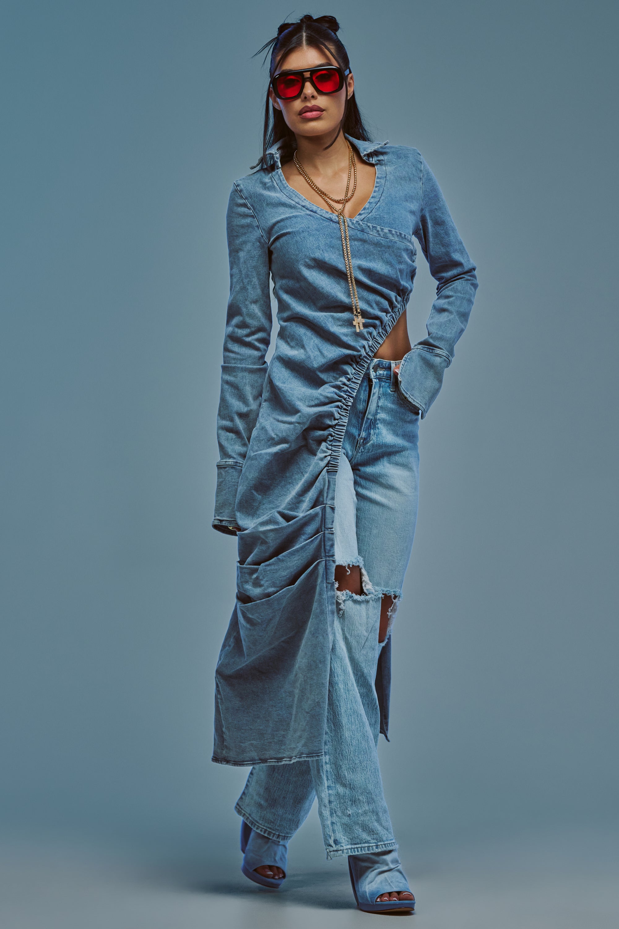 Street style is highlighted by a person wearing the GET MY CLOSE UP LONG DENIM BLOUSE styled as a dress with a ruched drape, layered over ripped jeans, paired with blue heeled sandals, red sunglasses, and layered necklaces while standing against a blue background.