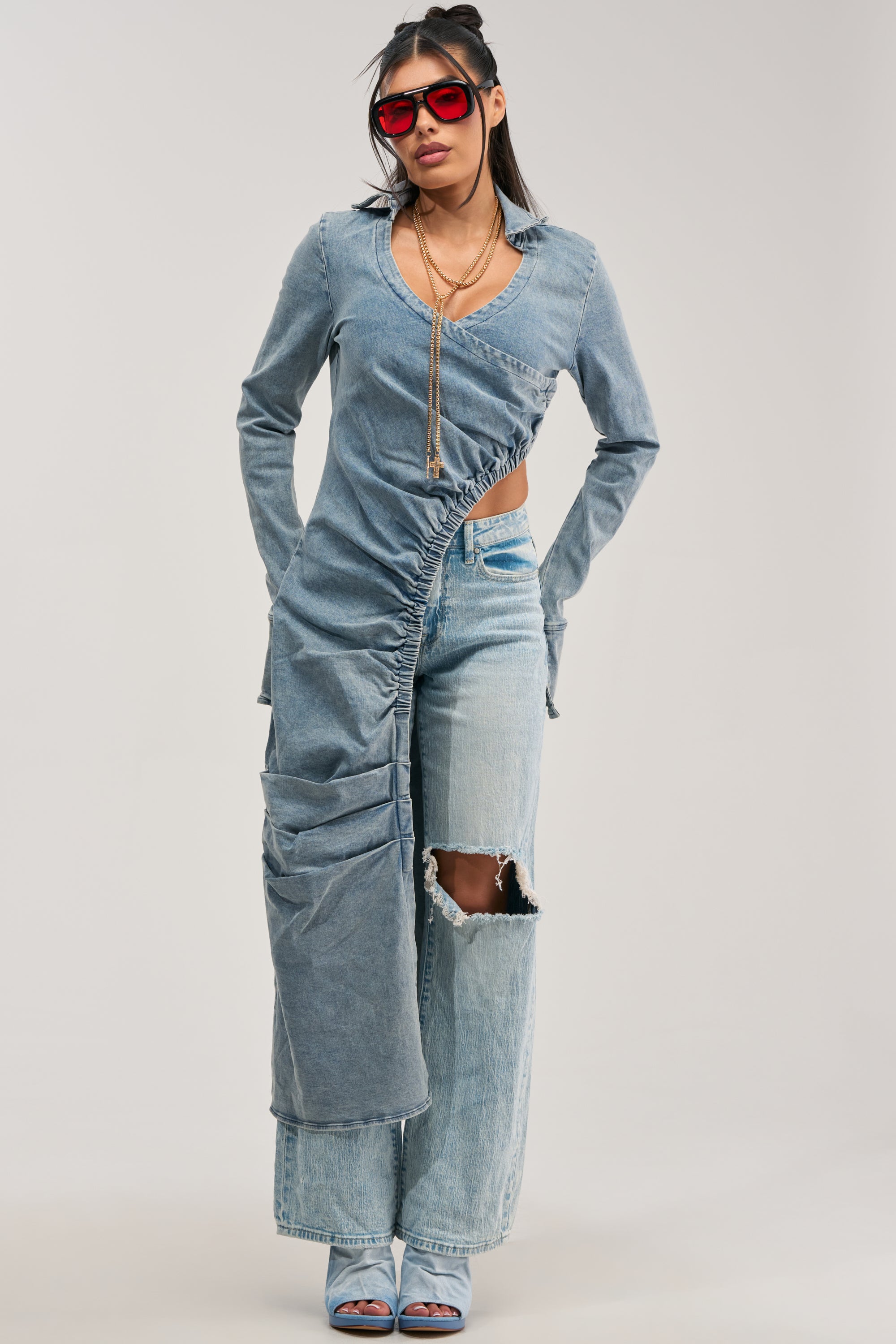 Wearing the GET MY CLOSE UP LONG DENIM BLOUSE styled asymmetrically as a dress over light-wash, knee-rip jeans, she completes her effortless street look with blue heeled sandals, red sunglasses, and layered necklaces while posing against a plain background.