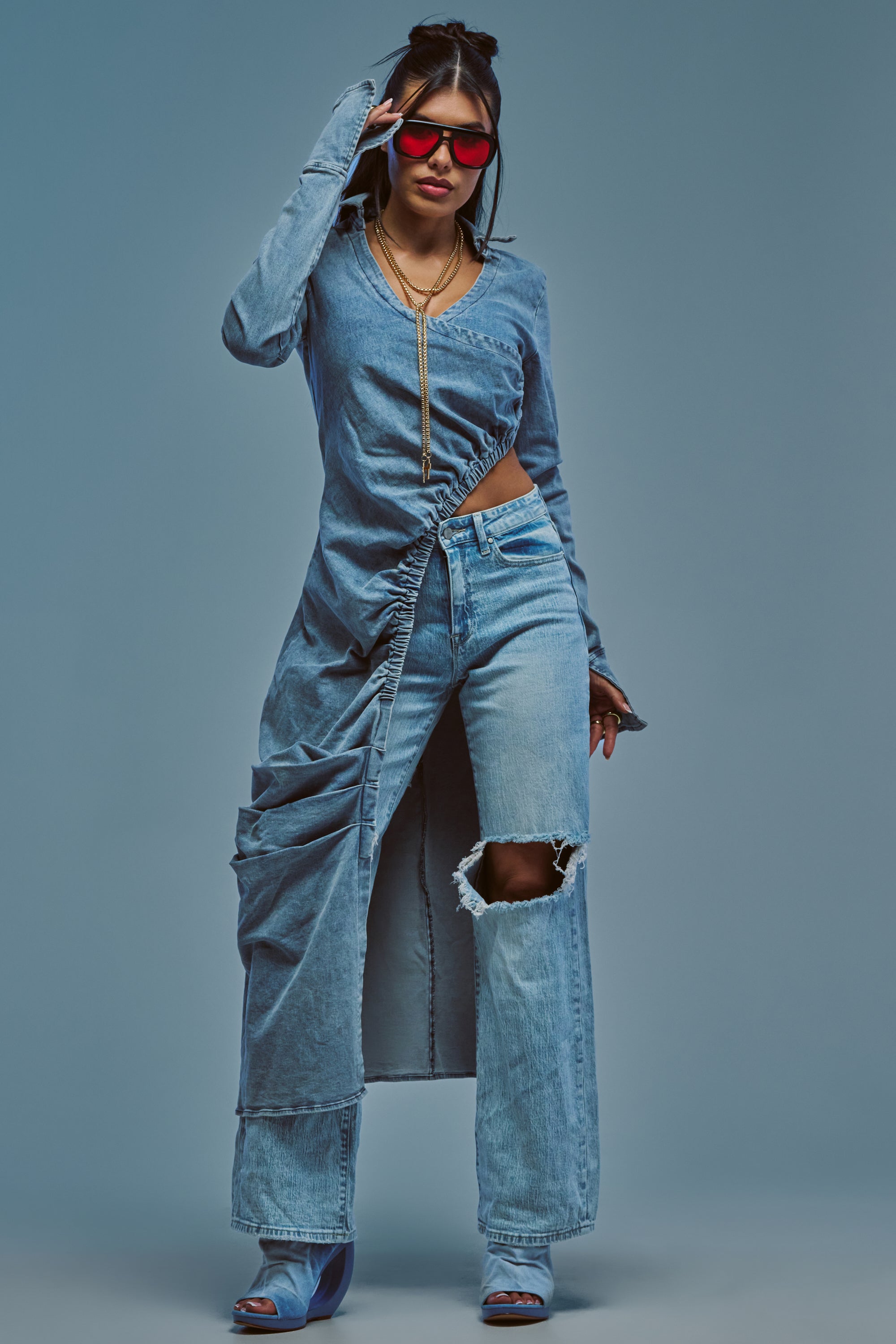 A woman wearing red sunglasses, ripped jeans, blue heels, and the GET MY CLOSE UP LONG DENIM BLOUSE stands confidently against a plain blue background.