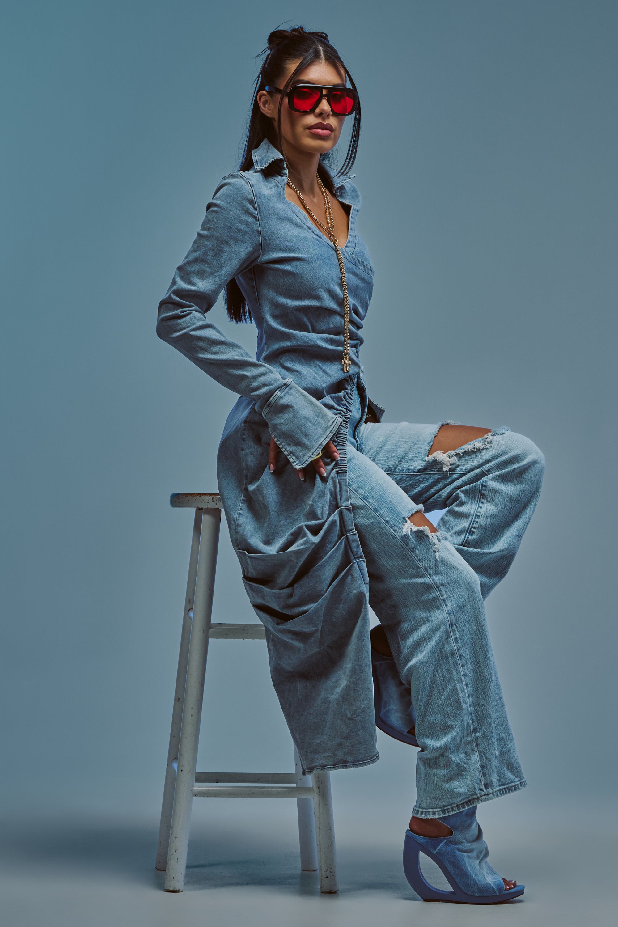 A woman in a long denim coat, ripped jeans, and blue open-toe heels sits on a stool against a blue background, wearing large red sunglasses and showcasing bold street style. Her look is completed with the GET MY CLOSE UP LONG DENIM BLOUSE, an asymmetrical wrap piece that adds a modern twist to her ensemble.