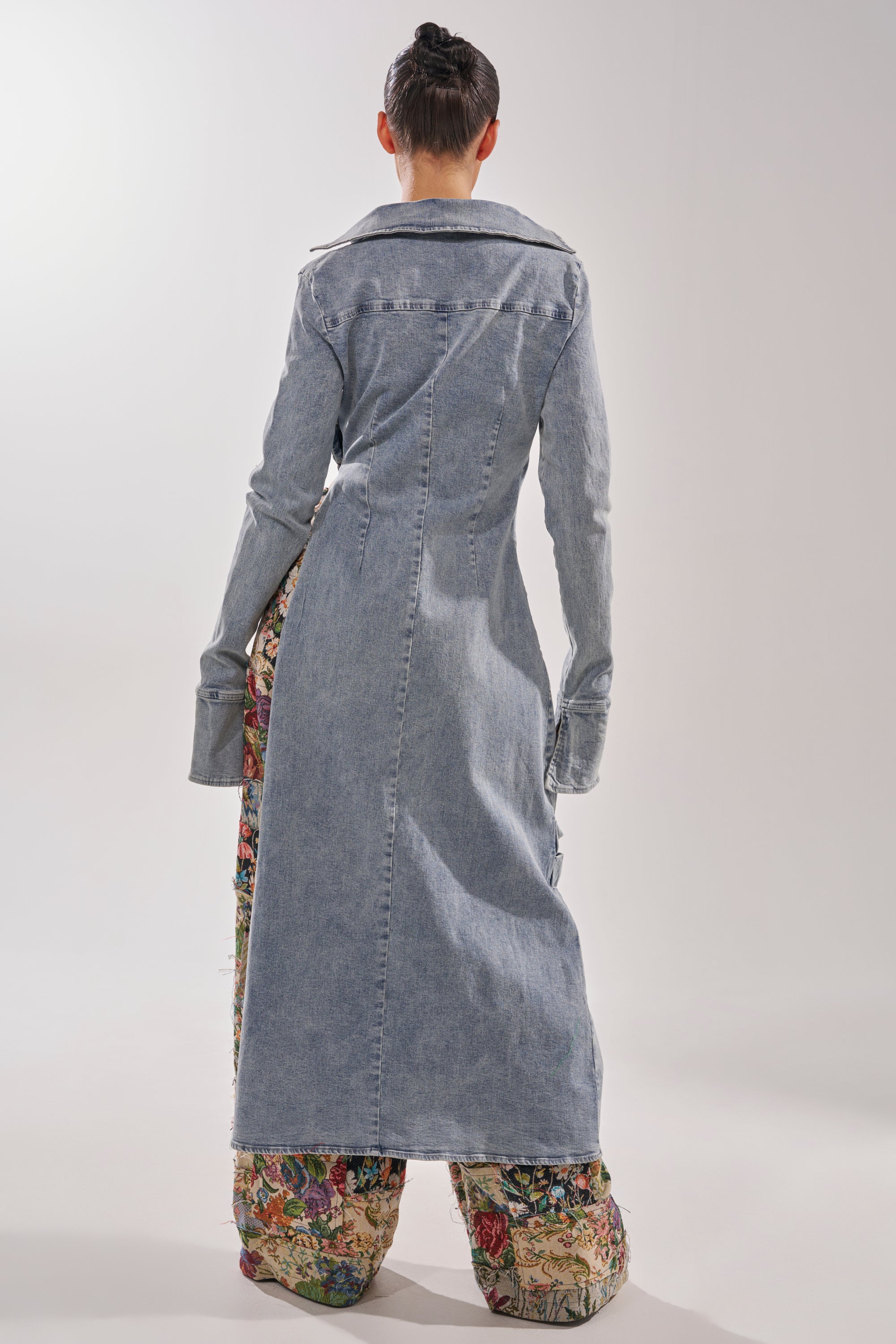 A person stands facing away in the GET MY CLOSE UP EMBELLISHED BUTTON DOWN SHIRT, a long light blue denim top with wide cuffs, paired with loose floral-print pants. Their hair is in a neat bun and the softly lit background is plain. 4