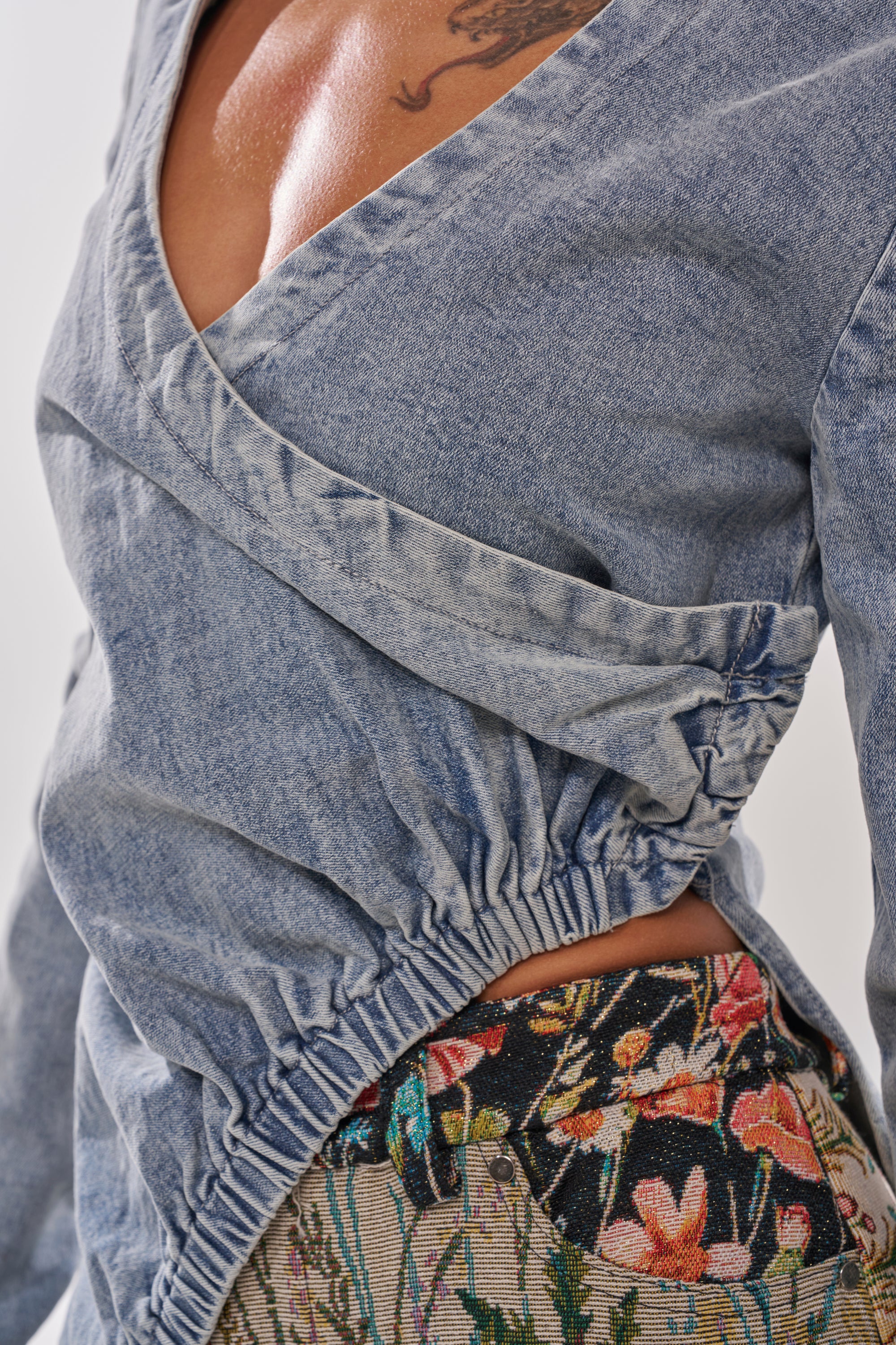 Close-up of a person wearing the GET MY CLOSE UP EMBELLISHED BUTTON DOWN SHIRT in light blue denim with a deep V-neck and ruched waist, paired with floral-patterned pants. A tattoo on their upper chest is partially visible. 5