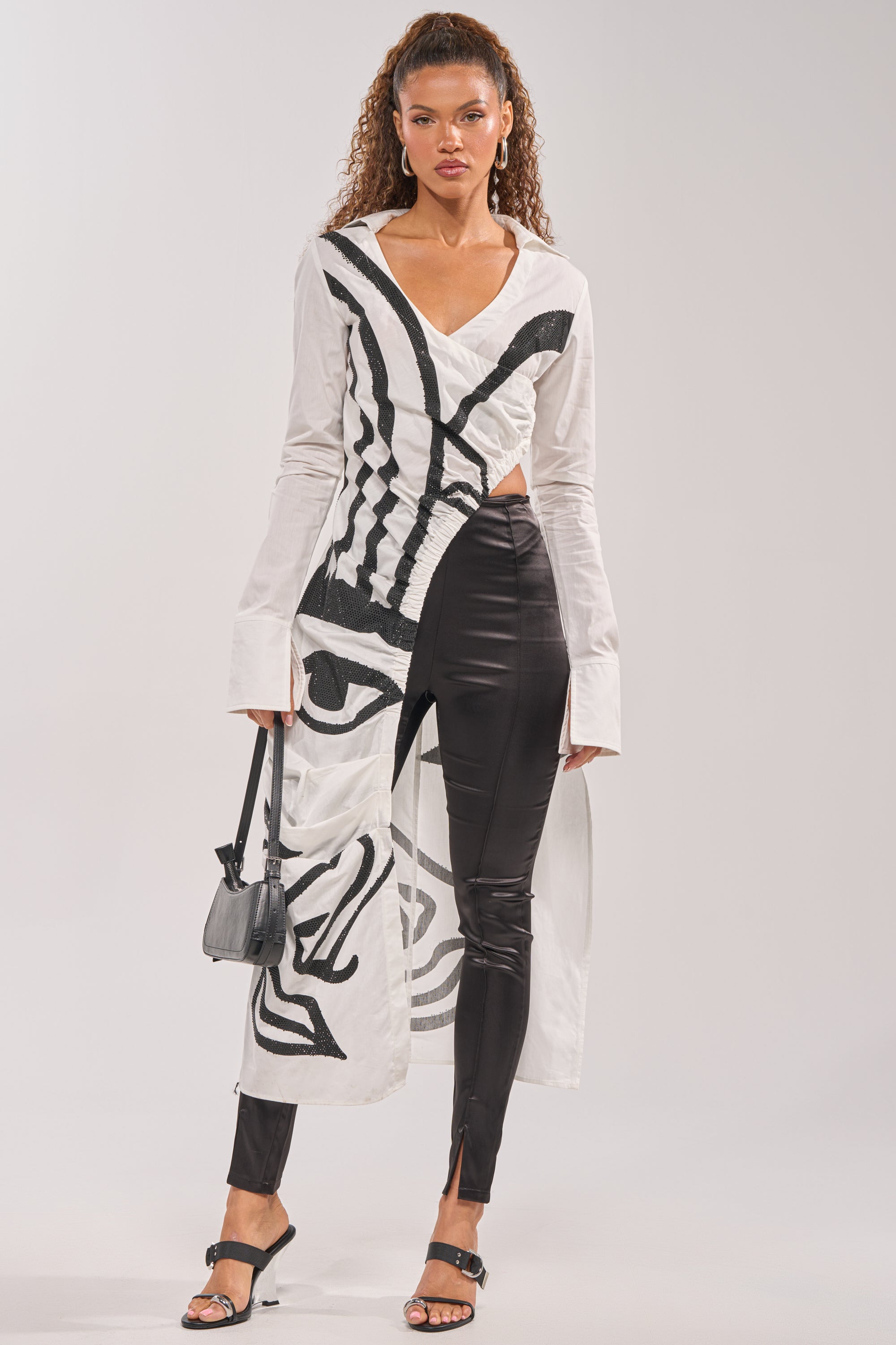 A woman with curly hair pulled back poses in the GET MY CLOSE UP EMBELLISHED BUTTON DOWN SHIRT—a long white top with bold black abstract face art—paired with black shiny fitted pants, sandals, and a small black handbag.