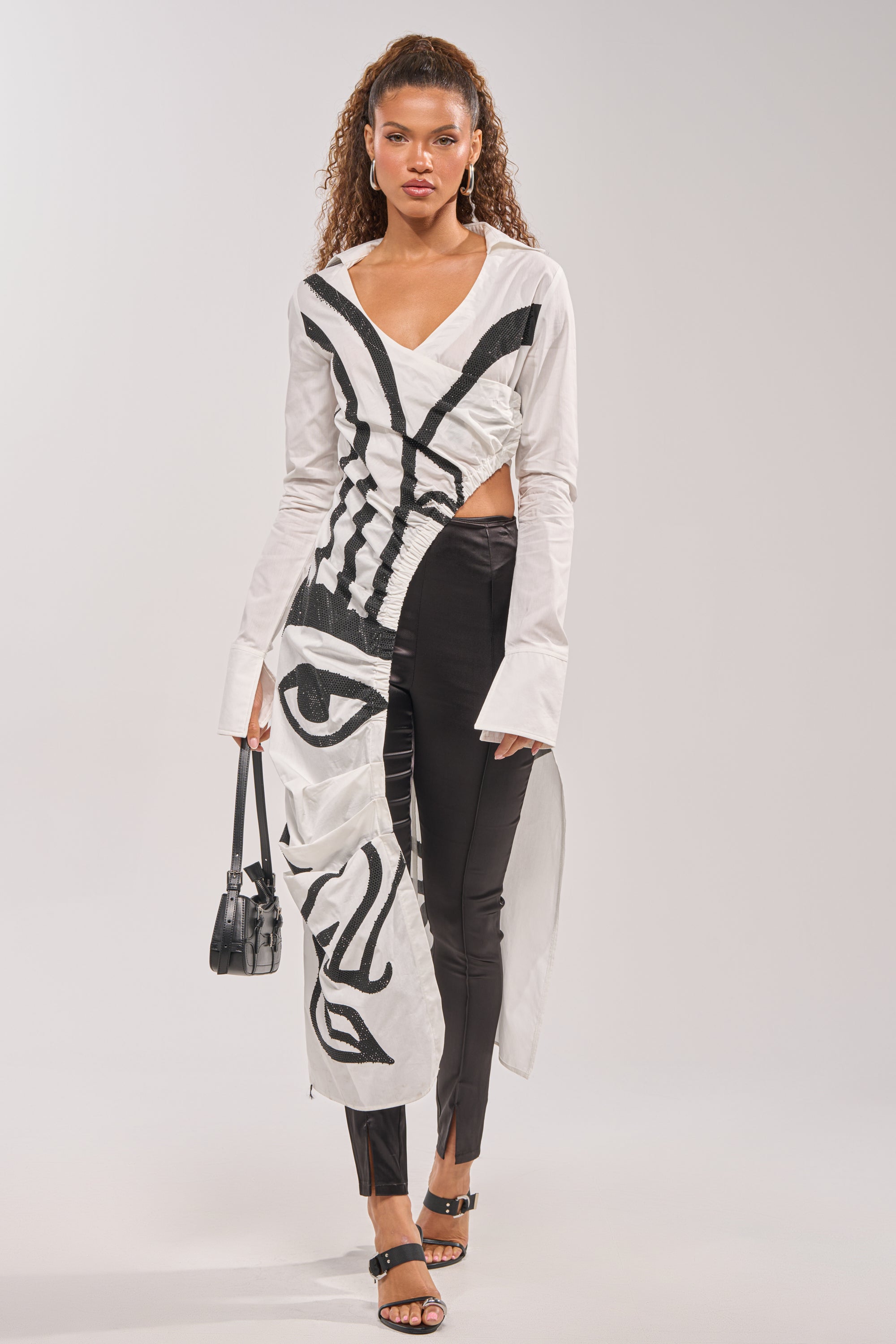 A woman wears the GET MY CLOSE UP EMBELLISHED BUTTON DOWN SHIRT, featuring an asymmetrical white poplin design with a black abstract face print and long sleeves—styled with black pants and a half-up ponytail for a bold date night look.