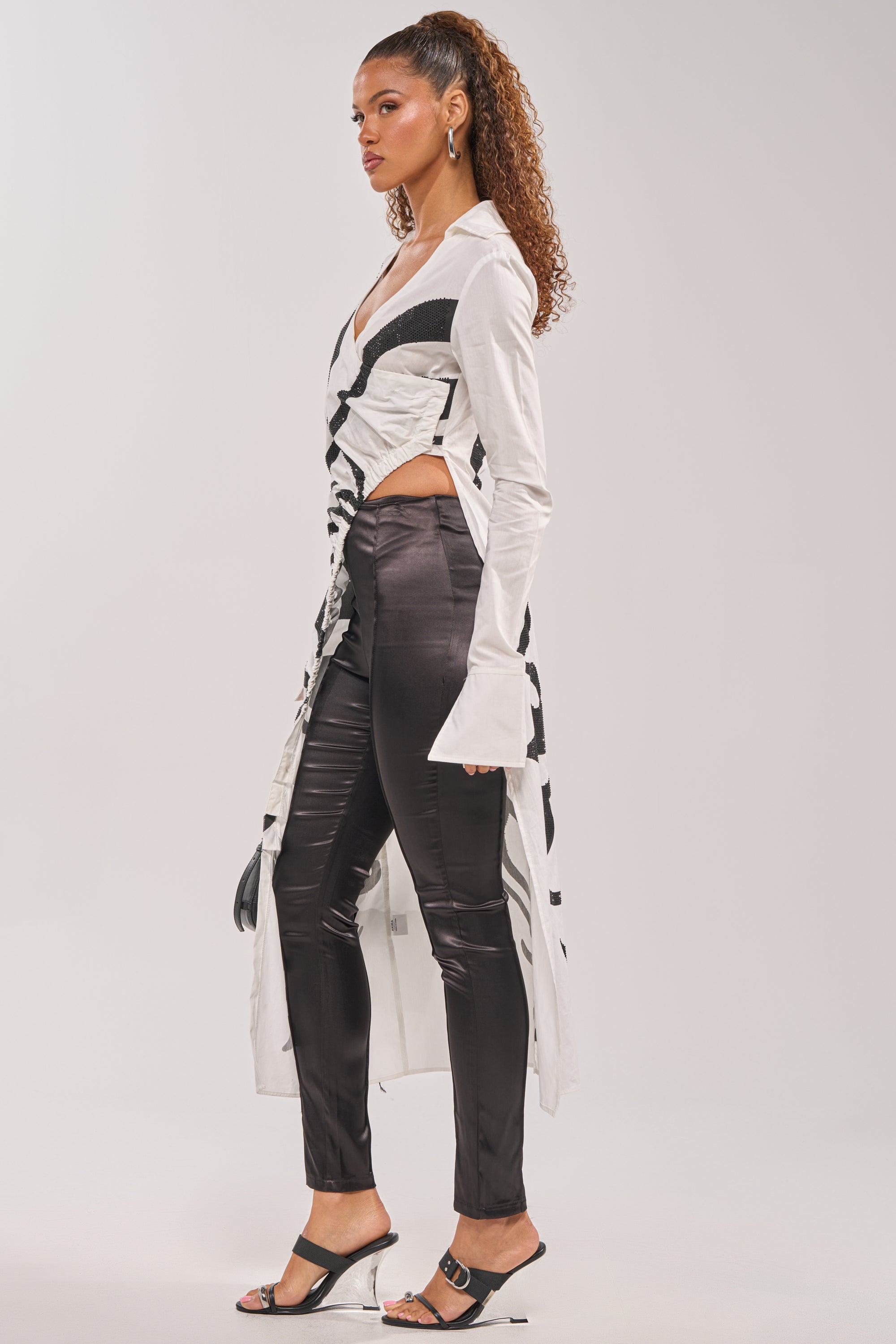 A woman poses confidently in profile against a plain background, wearing the GET MY CLOSE UP EMBELLISHED BUTTON DOWN SHIRT with black patterned details, shiny black pants, heeled sandals, hoop earrings, and her curly hair pulled back.