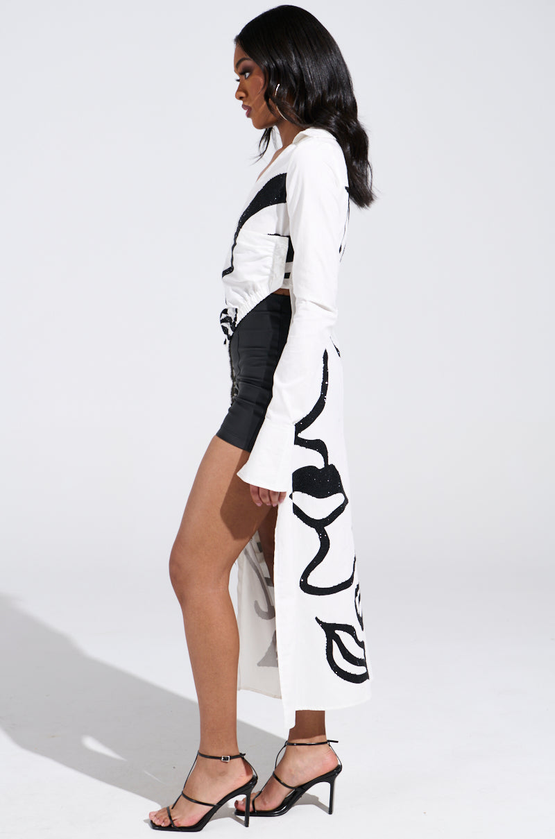 A person stands in profile against a plain backdrop, sporting the GET MY CLOSE UP EMBELLISHED BUTTON DOWN SHIRT—a white long-sleeved garment with bold black abstract patterns—paired with a black mini skirt and open-toe heels. Their long, wavy hair completes this chic look.