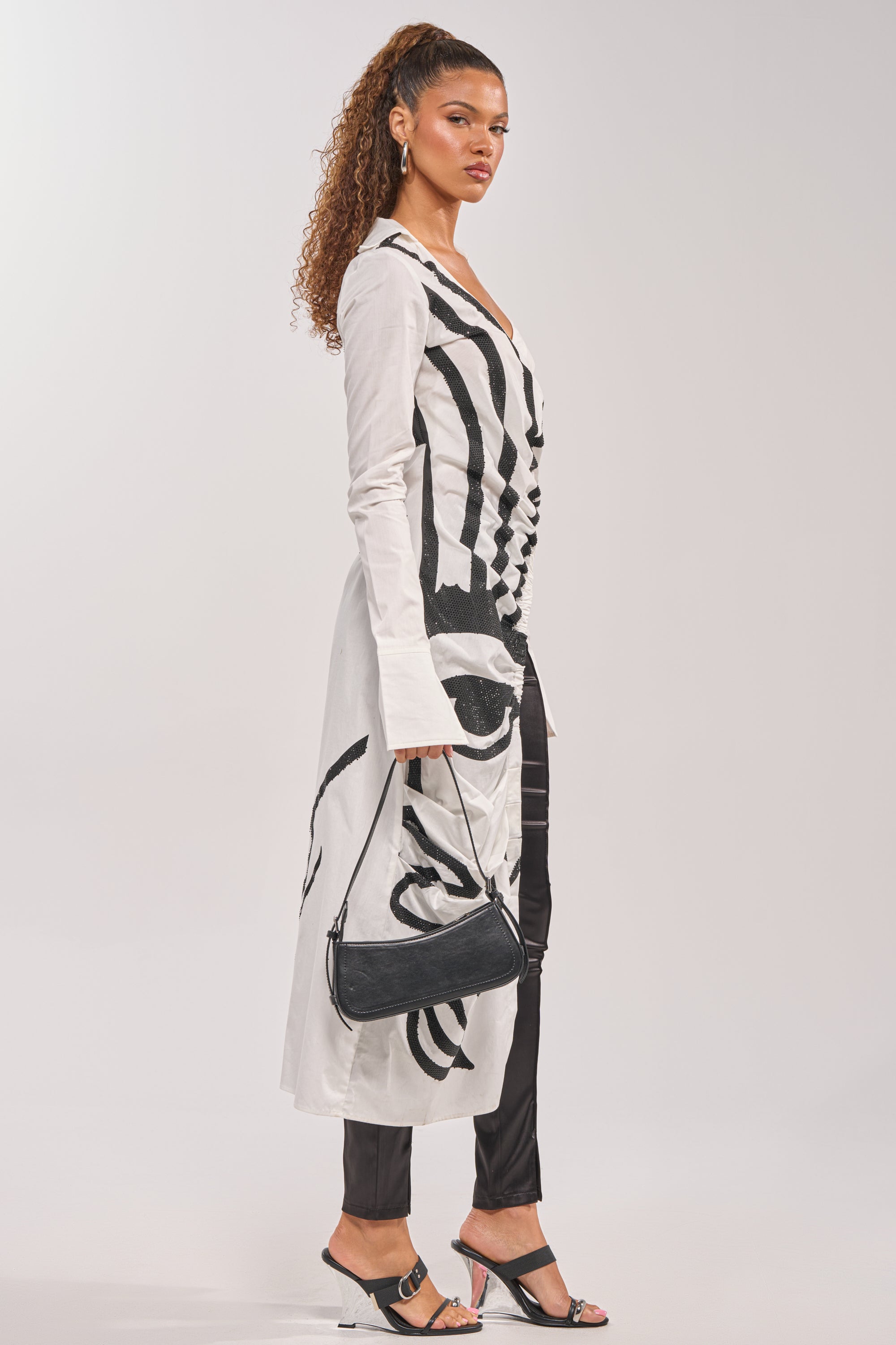 Model wearing the GET MY CLOSE UP EMBELLISHED ASYMMETRICAL BLOUSE, styled as a maxi shirt dress with black abstract print, paired with black fitted trousers, clear wedge heels, and holding a small black shoulder bag.