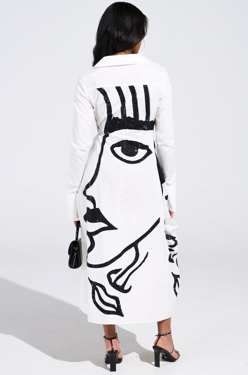 A person with long dark hair faces away, wearing the "GET MY CLOSE UP EMBELLISHED BUTTON DOWN SHIRT," a white coat with a bold abstract black face design. This statement piece is paired with a small black handbag and strappy black heels.