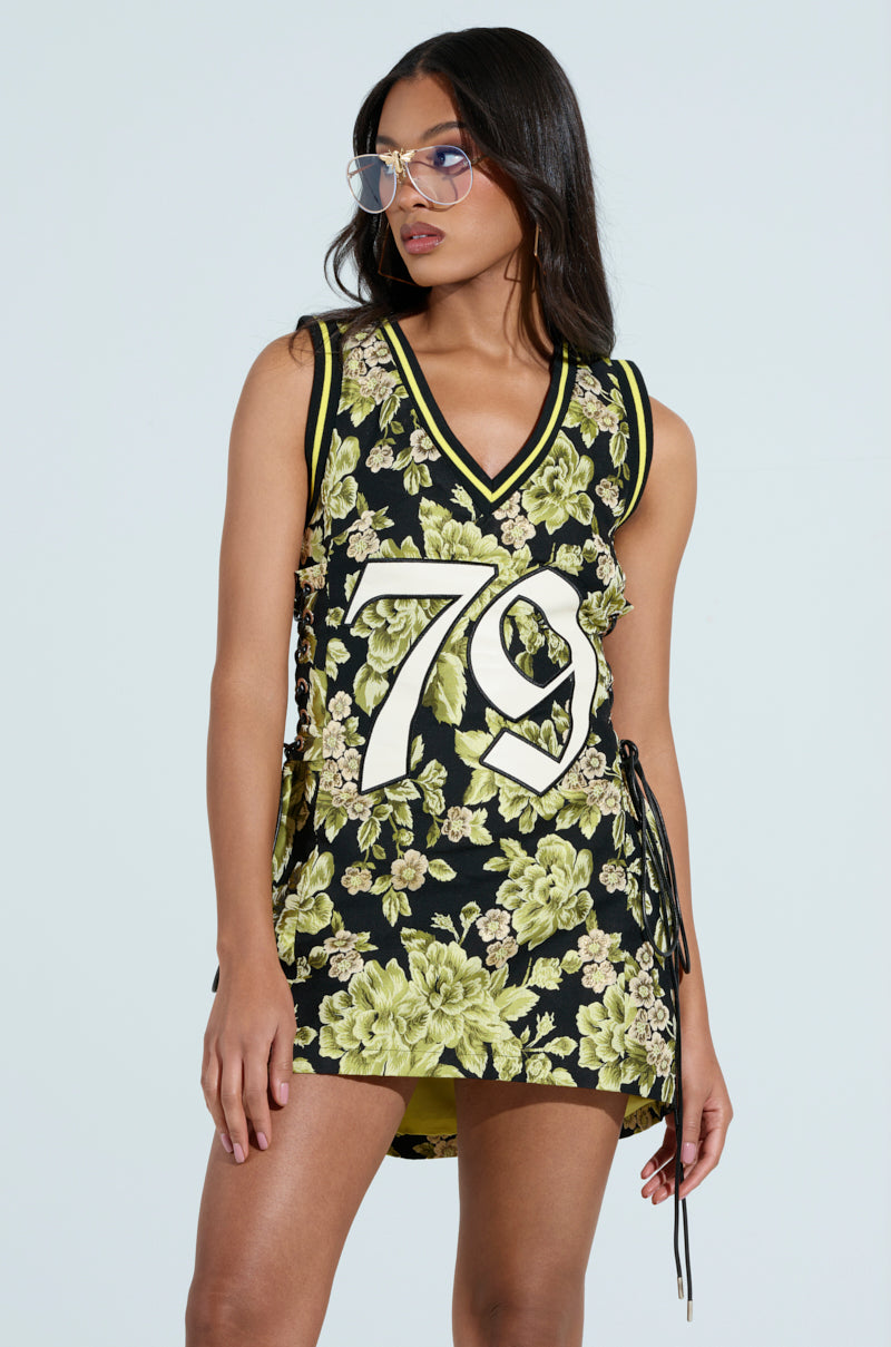 A woman in glasses wears the RENAISSANCE ON THE COURT BROCADE MINI DRESS, a sleeveless black dress with green floral patterns and white '79' on the front. Athletic accent stripes add a sporty edge as she gazes to the side with a neutral expression, hands relaxed by her side.