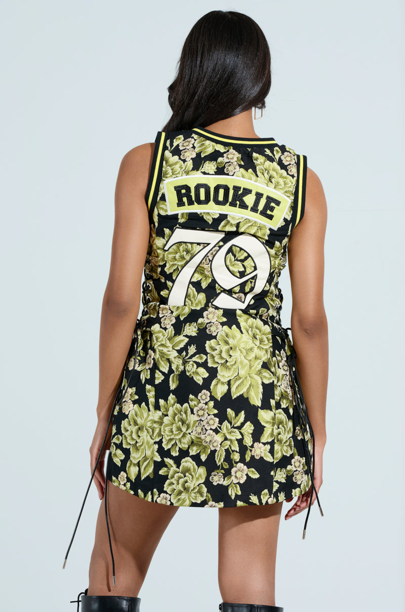 A person faces away, showcasing the "Renaissance on the Court" brocade mini dress adorned with vibrant floral patterns and "ROOKIE 70" on the back. The dress includes lace-up side panels and is stylishly paired with knee-high black boots.