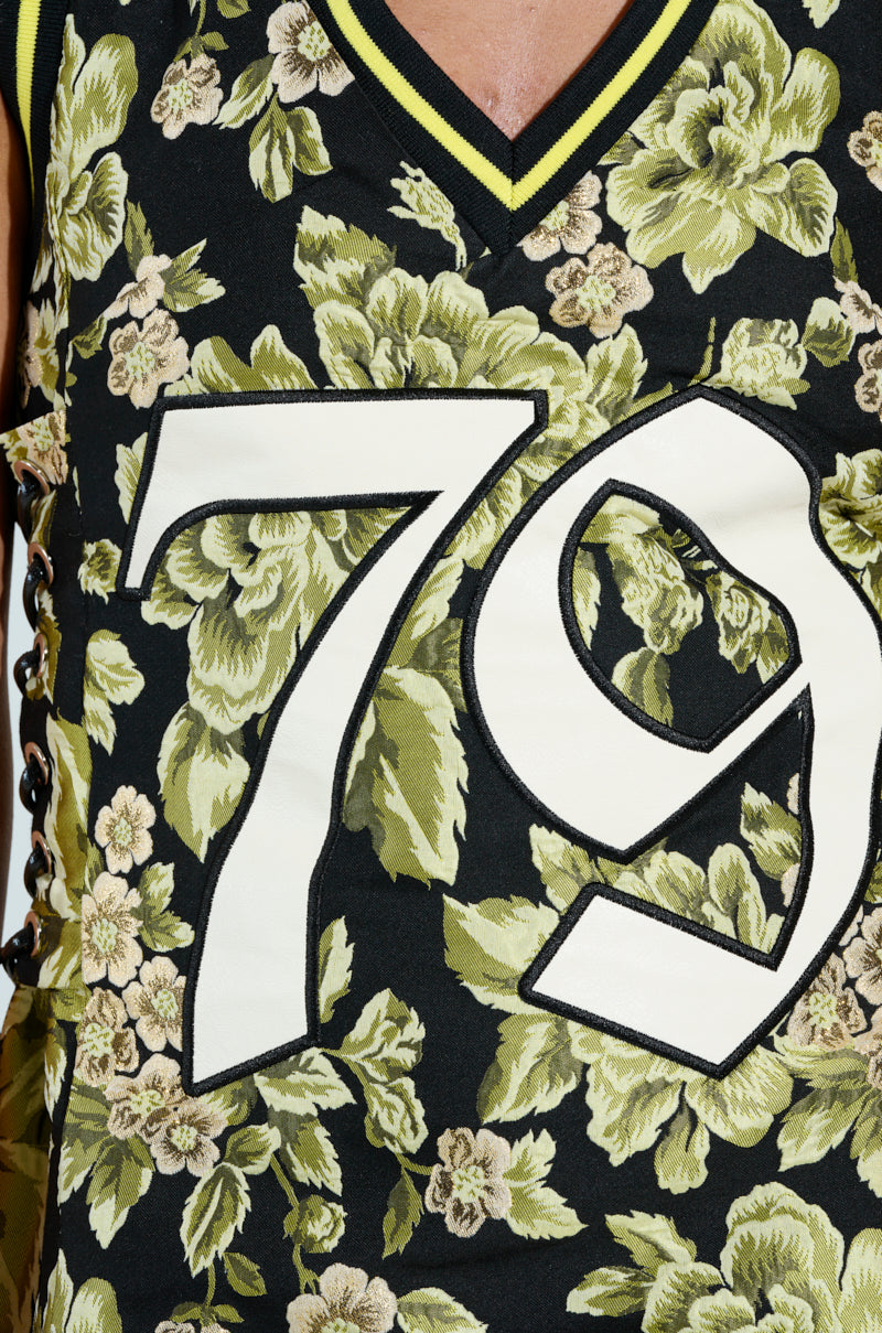 The RENAISSANCE ON THE COURT BROCADE MINI DRESS features a close-up of floral-patterned fabric, showcasing large light green and beige flowers. A white "79" with black stitching is embroidered on the sleeveless dress, which also includes athletic accent stripes on its yellow and black neckline.