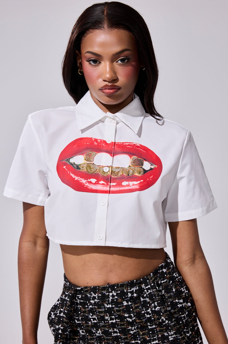 A woman models the NEW GRILLZ CROPPED BUTTON DOWN SHIRT, which features glossy red lips with rhinestone grills, paired with black high-waisted patterned pants against a plain background. 0