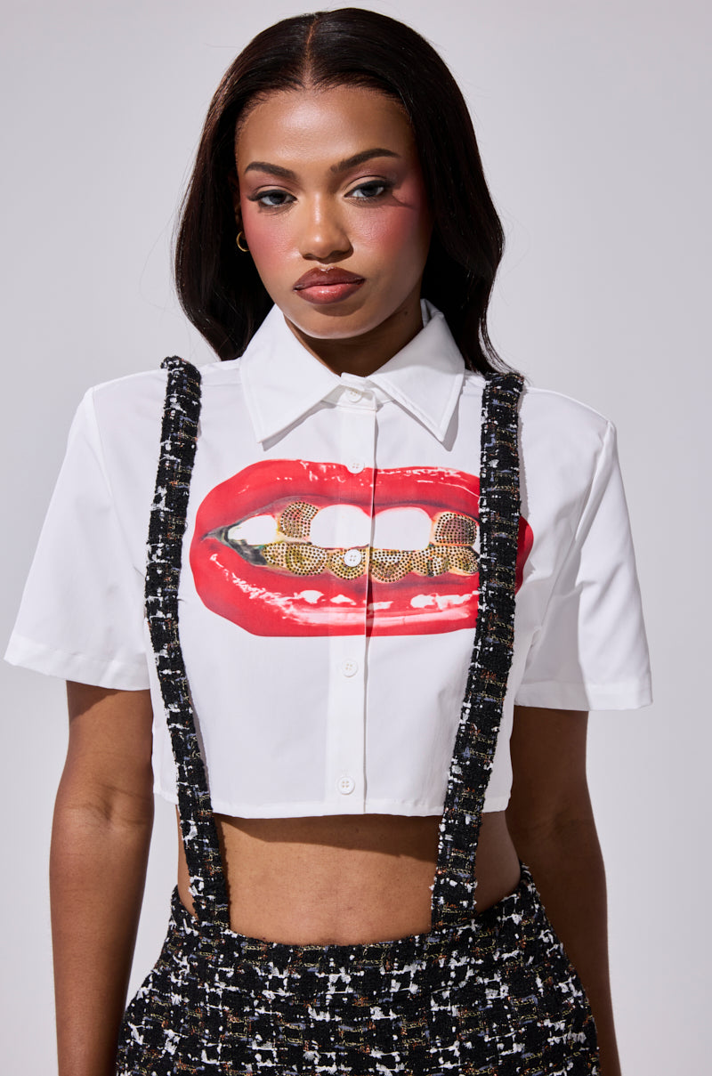 A woman with long dark hair rocks the NEW GRILLZ CROPPED BUTTON DOWN SHIRT, featuring red lips and rhinestone grill details, styled with high-waisted black-and-white textured suspenders and vibrant pink makeup. 1