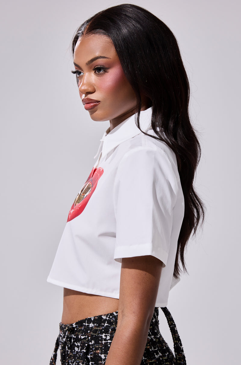 A woman with long, dark wavy hair stands in profile against a plain light background, wearing the NEW GRILLZ CROPPED BUTTON DOWN SHIRT—white, short-sleeved, cropped, with a red and gold front emblem. 2
