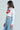 A person with long dark hair wears the LIPS BUTTON DOWN TOP, a white cropped poplin shirt with a bold lip graphic, paired with high-waisted light blue jeans featuring pearl-like embellishments, standing against a plain background.