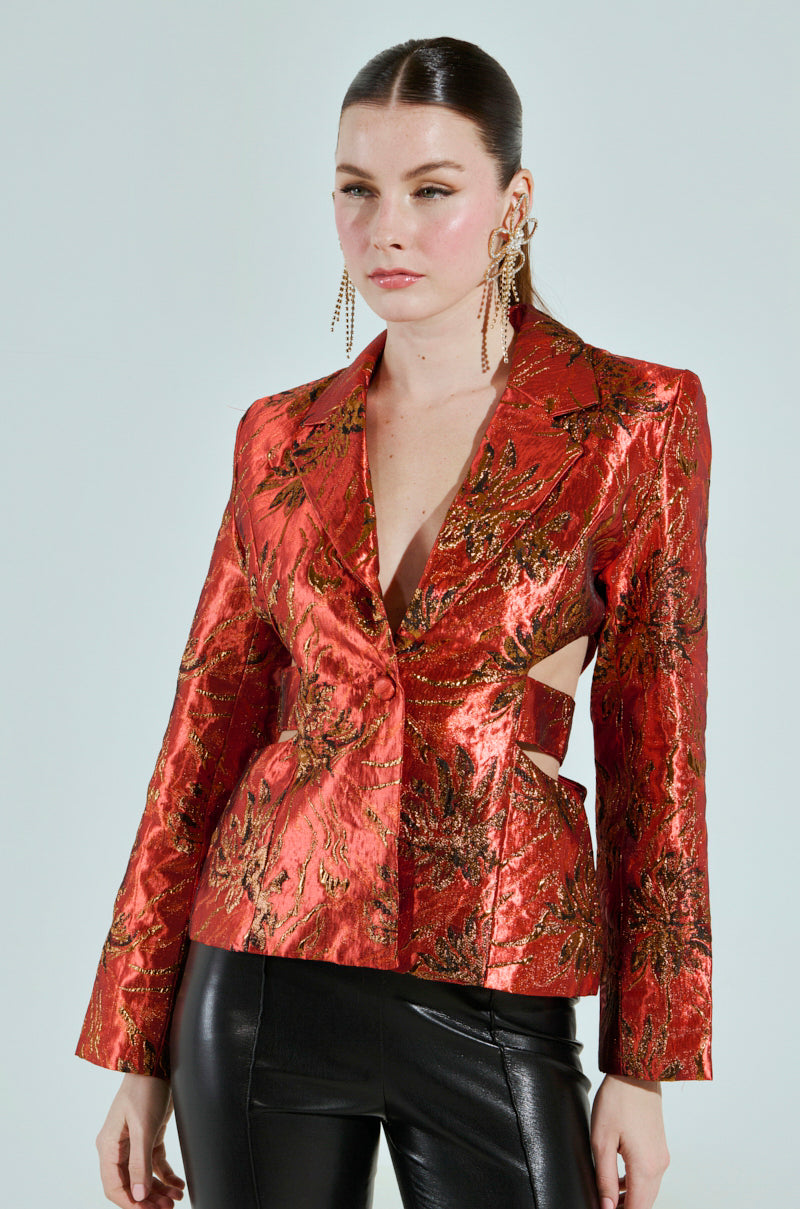 A person with long hair is wearing the shiny red COCKTAIL HOUR CUT-OUT BLAZER made from brocade and black leather pants, accessorized with long, dangling earrings while posing against a light gray backdrop.