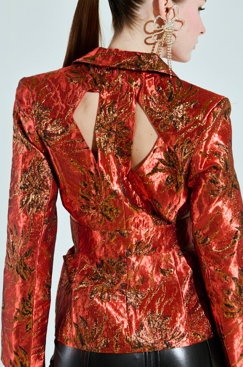 A person is wearing the COCKTAIL HOUR CUT-OUT BLAZER, a shiny red jacket with a metallic-infused pattern and brocade fabric, featuring unique cut-out designs on the back. They have long hair tied back and wear a distinctive large earring.