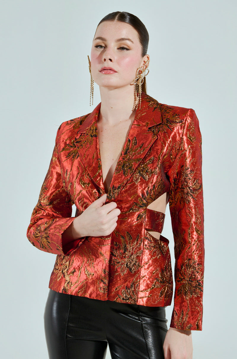 A woman wears the COCKTAIL HOUR CUT-OUT BLAZER in metallic red brocade with intricate sides, paired with black leather pants. Her hair is pulled back, and she sports statement gold earrings against a plain, minimal background.