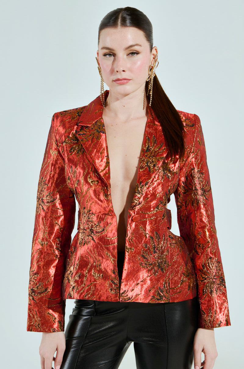 A person with long brown hair and gold earrings wears the COCKTAIL HOUR CUT-OUT BLAZER, a shiny red and gold metallic brocade jacket with a deep neckline, paired with black leather pants, against a light gray background.