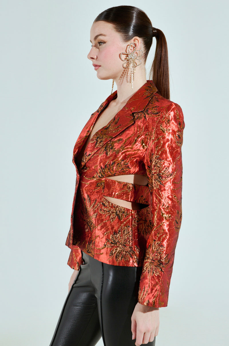 A woman is seen in profile wearing the COCKTAIL HOUR CUT-OUT BLAZER, made of shiny red brocade fabric, paired with black leather pants. She sports a sleek ponytail and large, elaborate earrings against a light gray background.