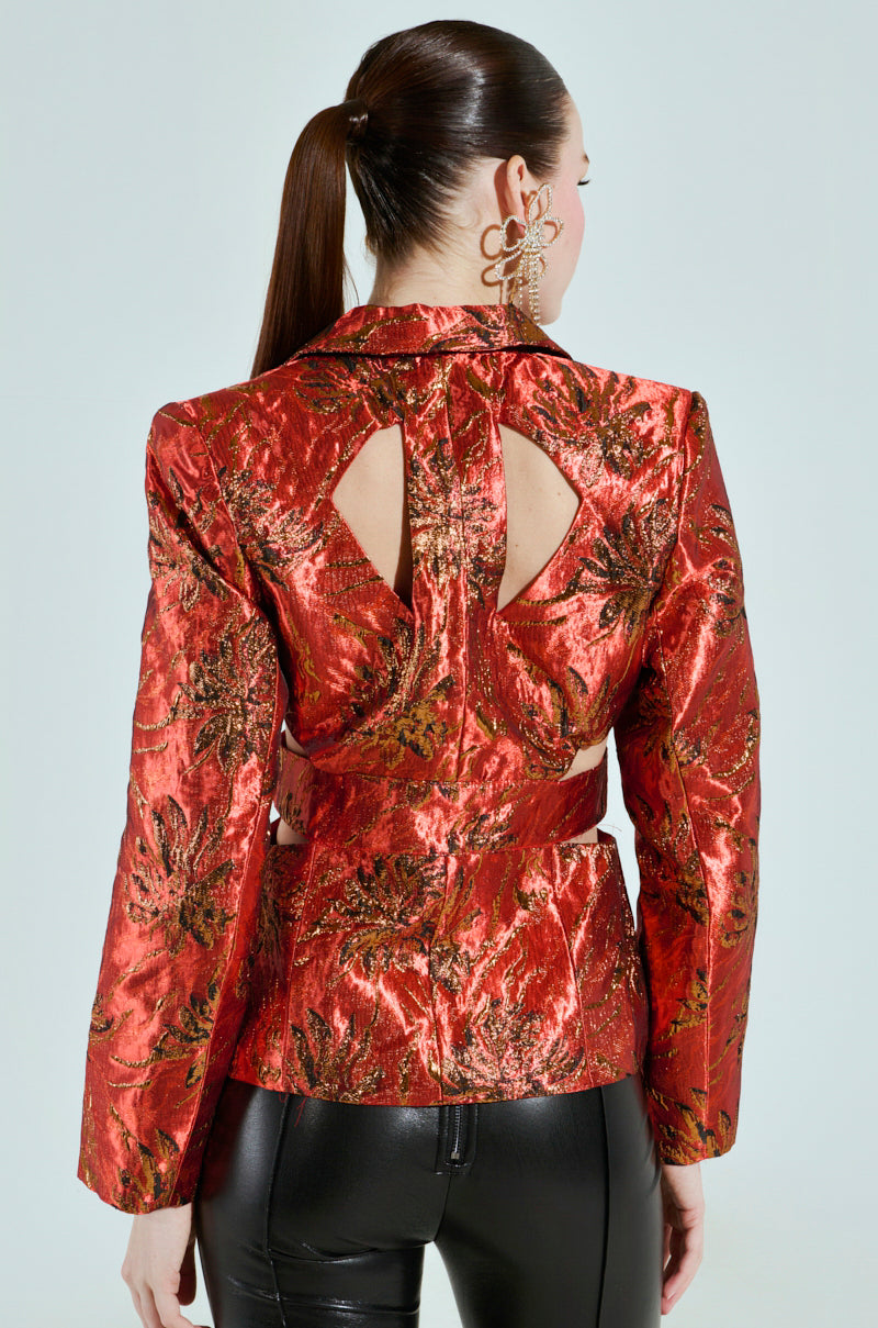 A person with long brown hair in a ponytail is wearing the red and gold COCKTAIL HOUR CUT-OUT BLAZER with intricate brocade fabric and triangular back cutouts, paired with shiny black pants. They are shown from behind against a light background.
