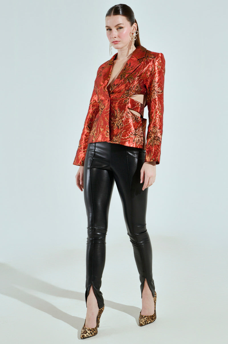 A woman confidently poses against a plain backdrop wearing the Cocktail Hour Cut-Out Blazer, featuring a shiny red brocade with metallic patterns and side cutouts, paired with black leather pants with ankle slits. Her look is completed with leopard print heels and statement earrings.