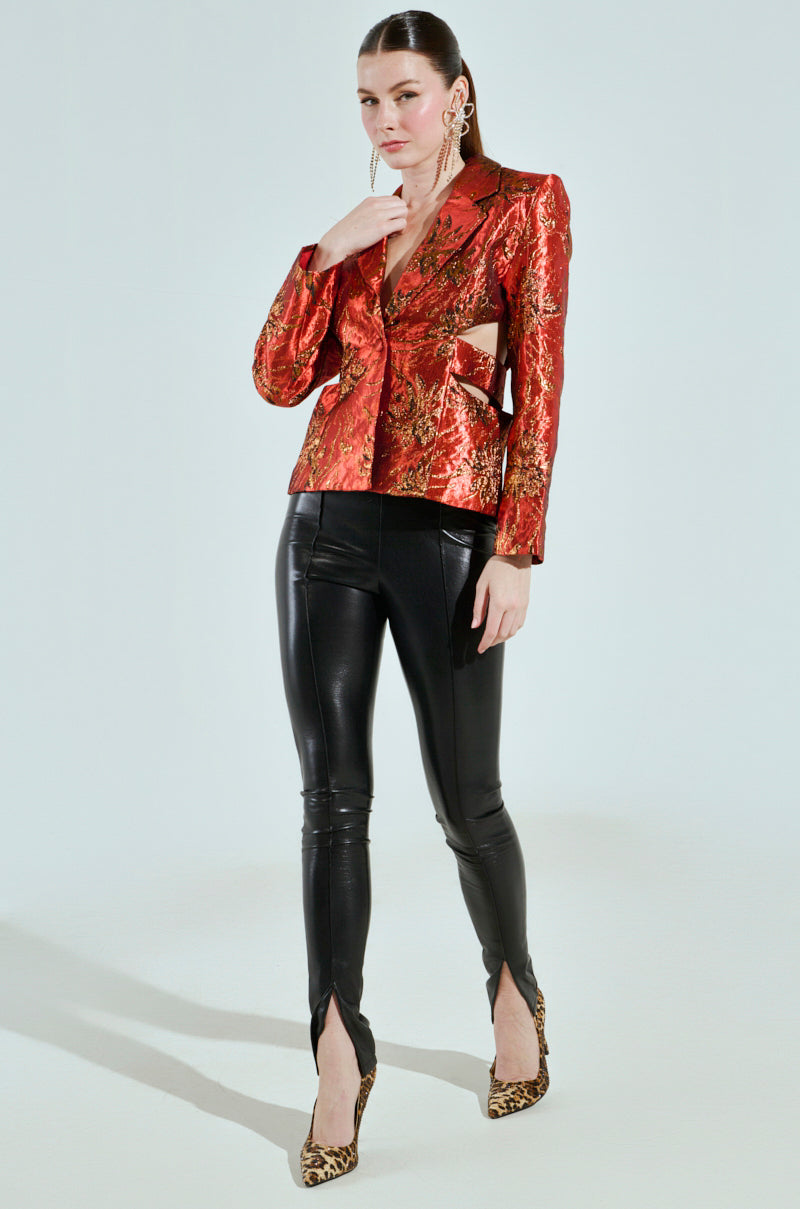 A woman poses in the COCKTAIL HOUR CUT-OUT BLAZER, featuring a red and gold metallic pattern with cutouts, paired with black leather leggings and leopard print heels. She stands against a light background, hand near her face to showcase her long earrings.