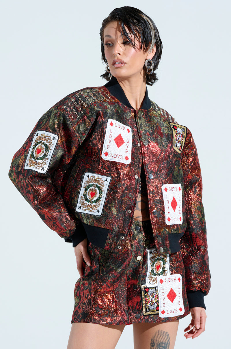 Someone is sporting the SHOW YOUR CARDS BROCADE BOMBER, featuring shiny playing card-inspired patches, paired with a skirt decorated with the queen of spades and king of clubs. The ensemble includes phrases like "Life & Love" and is completed with short, wet-look hair.
