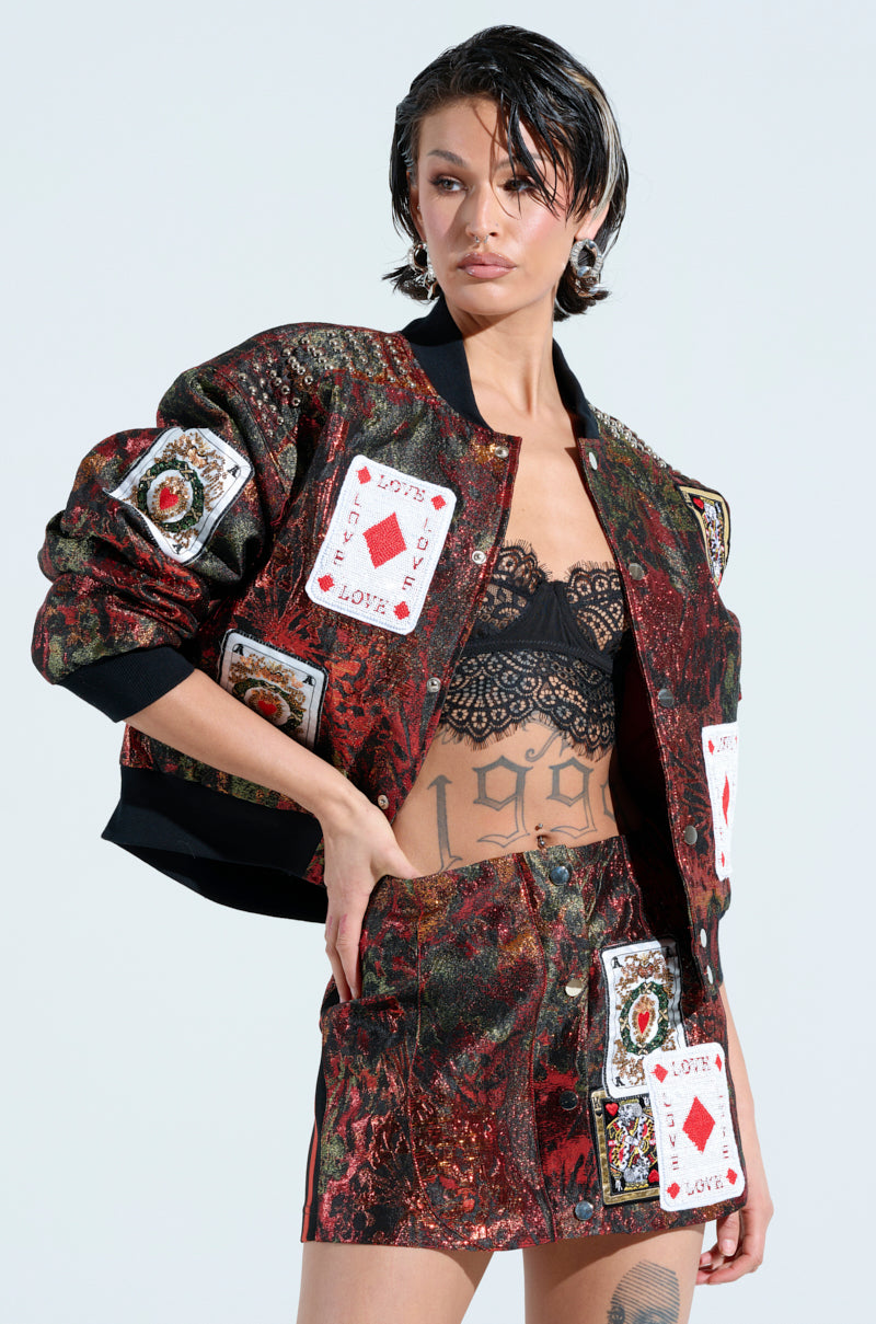 A model confidently showcases the SHOW YOUR CARDS BROCADE BOMBER, featuring playing card-inspired patches. A black lace bralette adds allure beneath, and tattoos peek from their abdomen. The plain, light background highlights the bold creativity of the ensemble.
