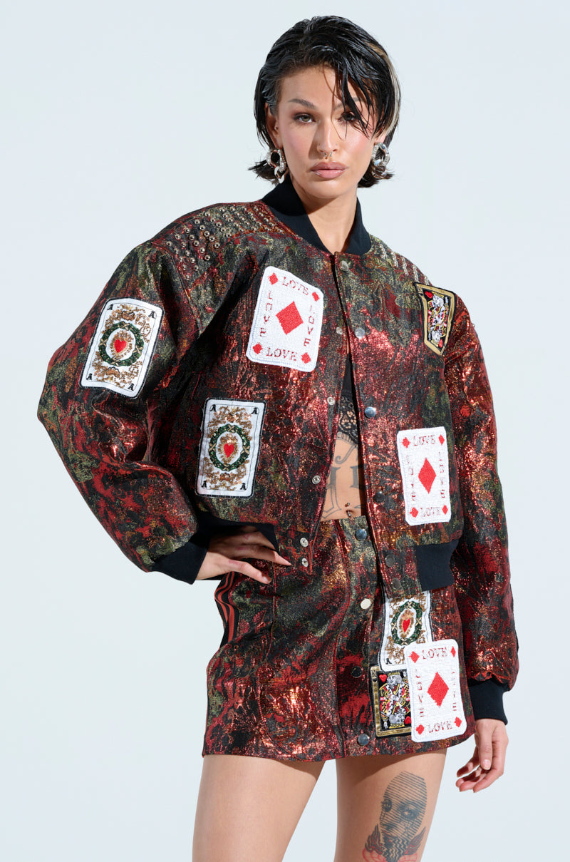 A person with short hair confidently poses with one hand on their hip, wearing the studded shoulder SHOW YOUR CARDS BROCADE BOMBER and a matching patterned skirt adorned with playing card patches, all set against a light, neutral background.