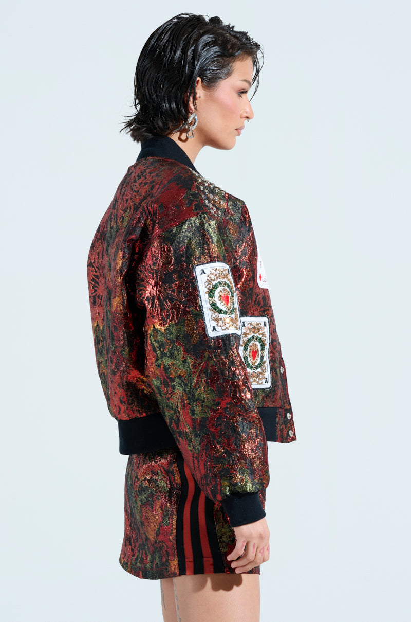 Side view of a person with short dark hair wearing the SHOW YOUR CARDS BROCADE BOMBER and a matching skirt, featuring playing card-inspired patches on the sleeves and a floral pattern. The background is plain and light-colored.