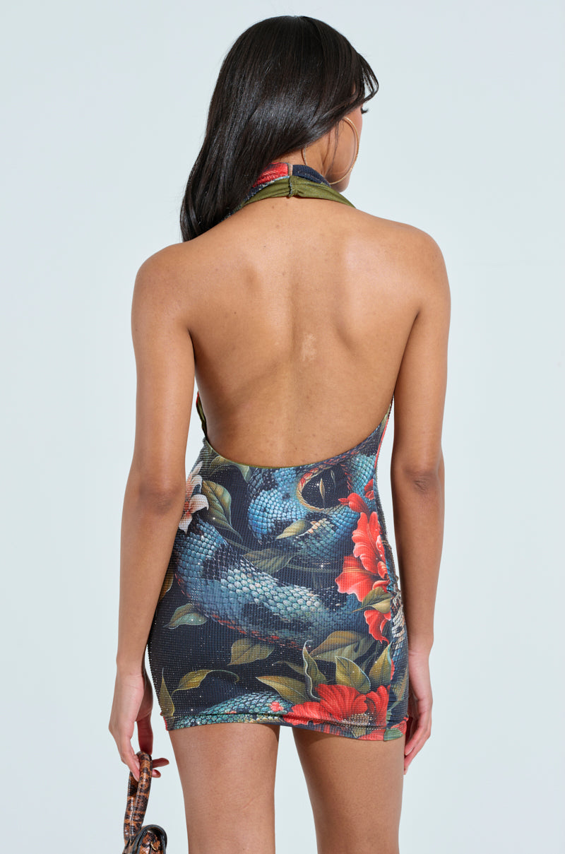 A woman with long dark hair models the SHOW STOPPER PRINTED RHINESTONE MINI DRESS, a sleeveless halter design featuring vivid floral and snake print. She holds a purse in her left hand against a light blue background, showcasing her jungle-inspired flair.
