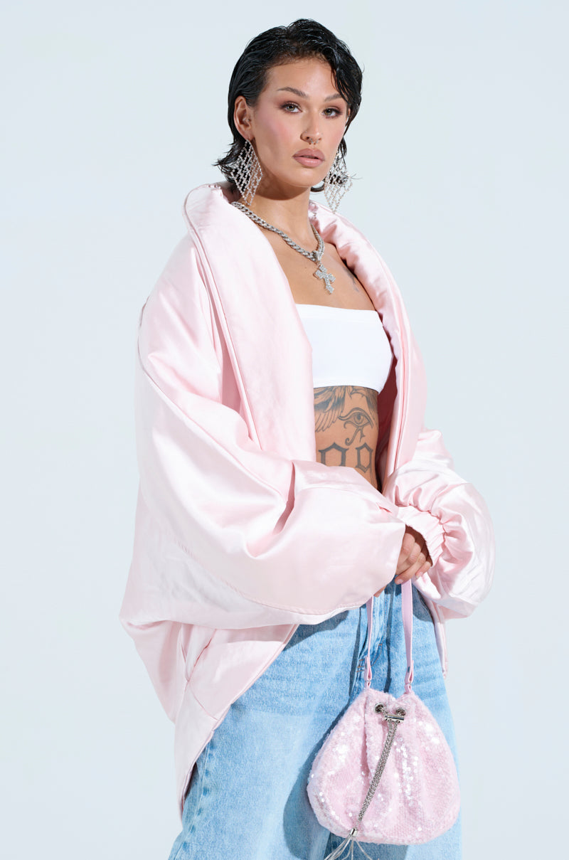 A person with short dark hair stands against a light background, wearing the FIRST CLASS COCOON COAT in pink, a white tube top, light blue jeans, and holding a pink sequined bag. They accessorize with statement earrings, a cross necklace, and display a torso tattoo.