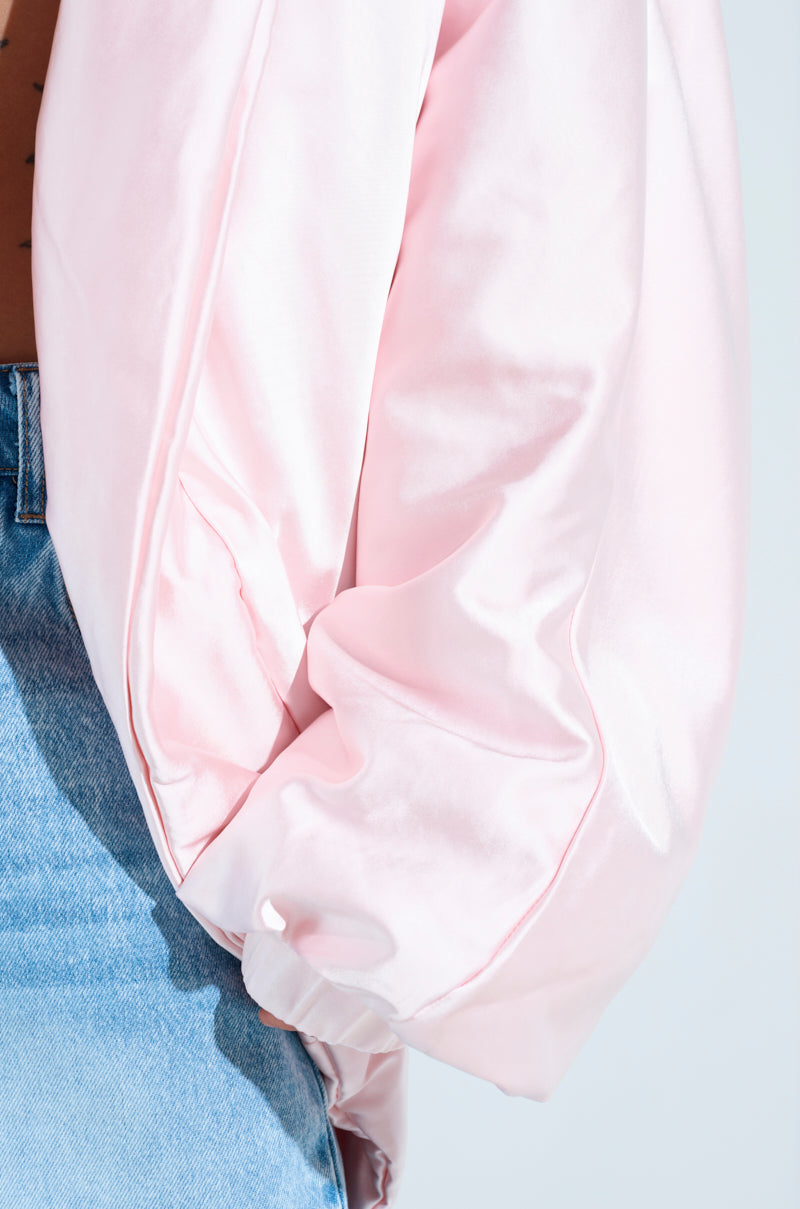 Close-up of a person in an oversized FIRST CLASS COCOON COAT with puffed sleeves, showcasing the shiny pink satin. It's paired with light blue denim jeans, highlighting the contrast between the soft pink fabric and rugged denim texture.