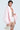 A person models the FIRST CLASS COCOON COAT in light pink, paired elegantly with a white netted dress and a pearl-detailed purse. The plain background accentuates their chic ensemble.
