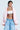 A person with short dark hair wears a pink FIRST CLASS COCOON COAT, a white tube top, and baggy blue jeans. Tattoos cover their chest and stomach. They accessorize with large earrings, a silver necklace, and hold a pink bag in one hand.