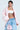 With short hair, they wear a pink FIRST CLASS COCOON COAT over a white bandeau top and light blue jeans, holding a sequined pink bag. A large 