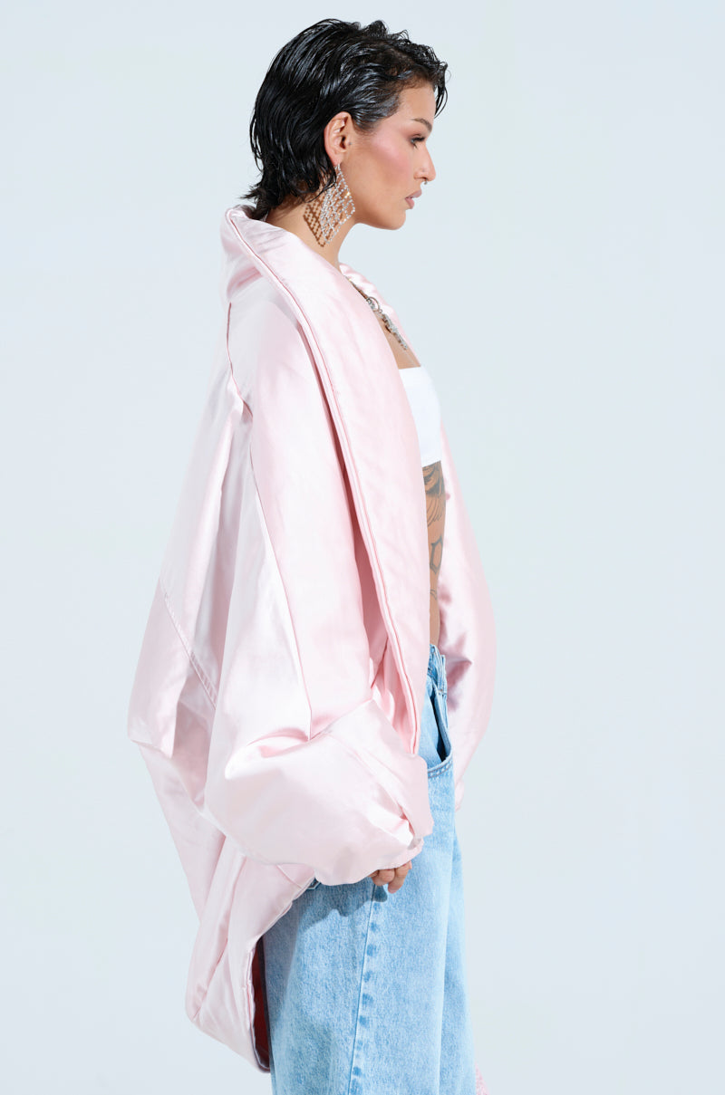 A person stands in profile against a white background, wearing the oversized FIRST CLASS COCOON COAT in light pink padded satin, along with a white tube top and blue jeans. They have short dark hair and are accessorized with large, intricate earrings.