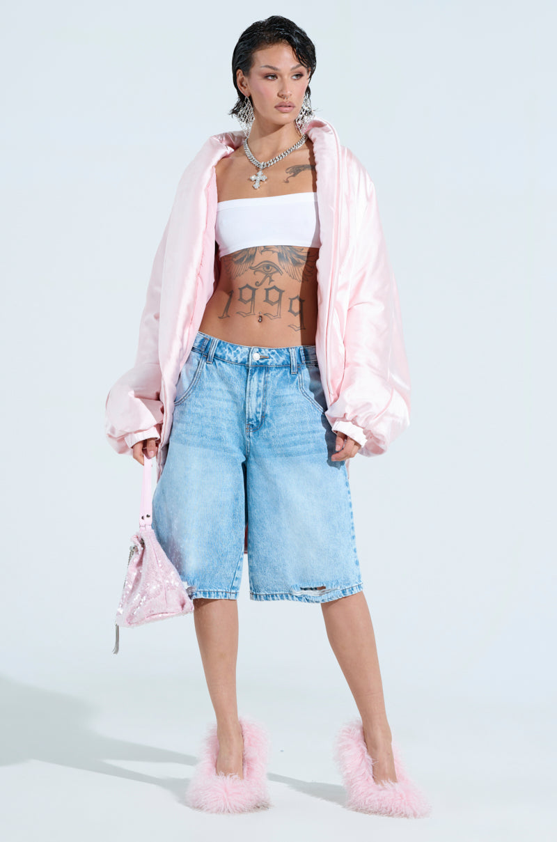Wearing an oversized FIRST CLASS COCOON COAT in pink, paired with a white tube top and denim shorts, this person stands confidently. They accessorize with a pink handbag, necklaces, and fluffy pink high-heeled slippers. Tattoos embellish their torso against the plain backdrop.