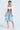 Wearing an oversized FIRST CLASS COCOON COAT in pink, paired with a white tube top and denim shorts, this person stands confidently. They accessorize with a pink handbag, necklaces, and fluffy pink high-heeled slippers. Tattoos embellish their torso against the plain backdrop.