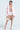 Sporting the FIRST CLASS COCOON COAT in light pink, a person confidently poses over a white netted dress and high-waisted bottoms. They accessorize with a beaded handbag and clear heels against a plain white background.