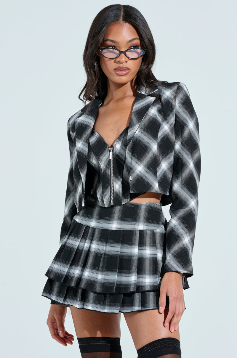 Wearing the DIRTY MARTINI CROPPED BLAZER with a matching pleated skirt and cropped top, a person exudes confidence. The satin-lined ensemble complements their long, dark hair and stylish eyeglasses, while a poised expression graces their face against the light background.