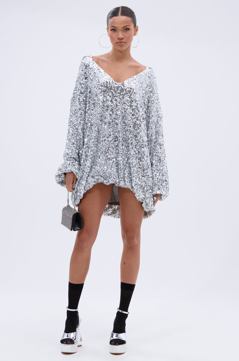 A woman exudes confidence in the GISELLE SEQUIN SWEATSHIRT DRESS IN SILVER, styled with a glittery handbag, platform sandals and black socks, large hoop earrings, and slicked-back hair for a bold party look.