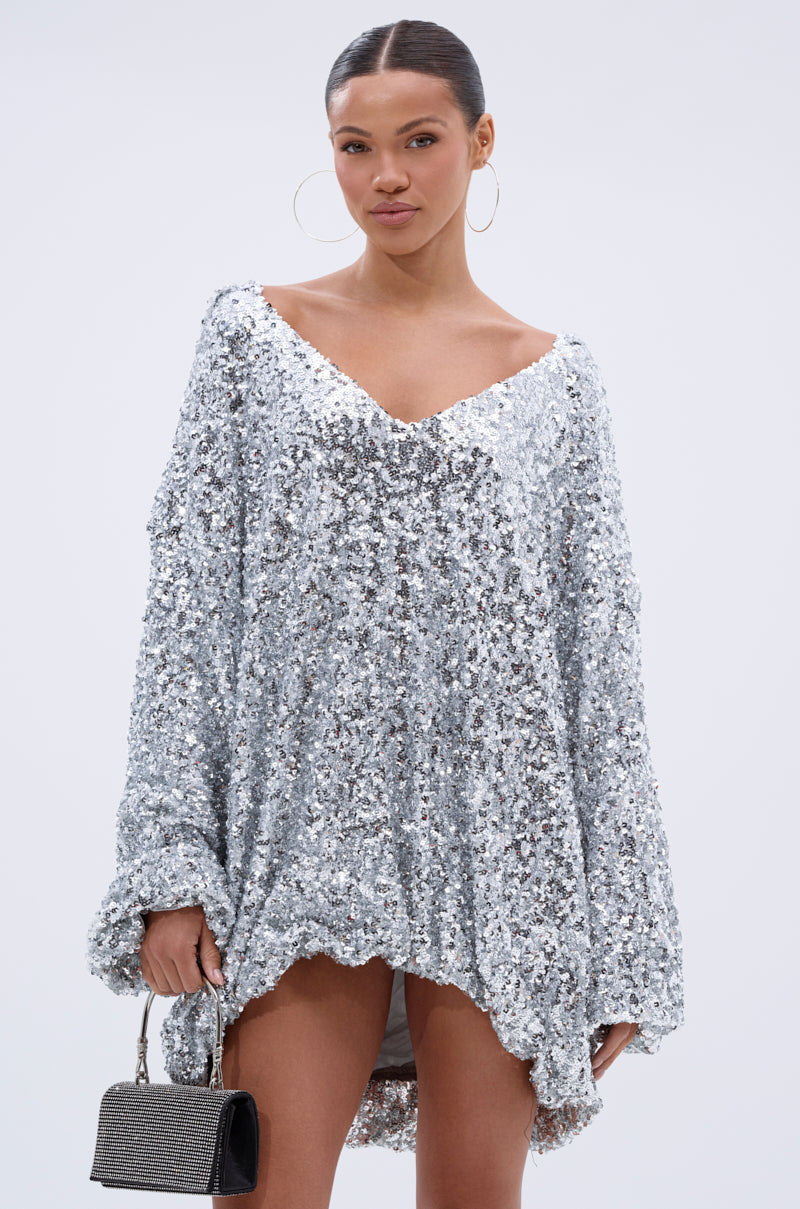 A woman models the GISELLE Sequin Sweatshirt Dress in Silver, featuring long sleeves and a deep V-neckline. She accessorizes with large hoop earrings and a small gray handbag, posing against a plain light background.