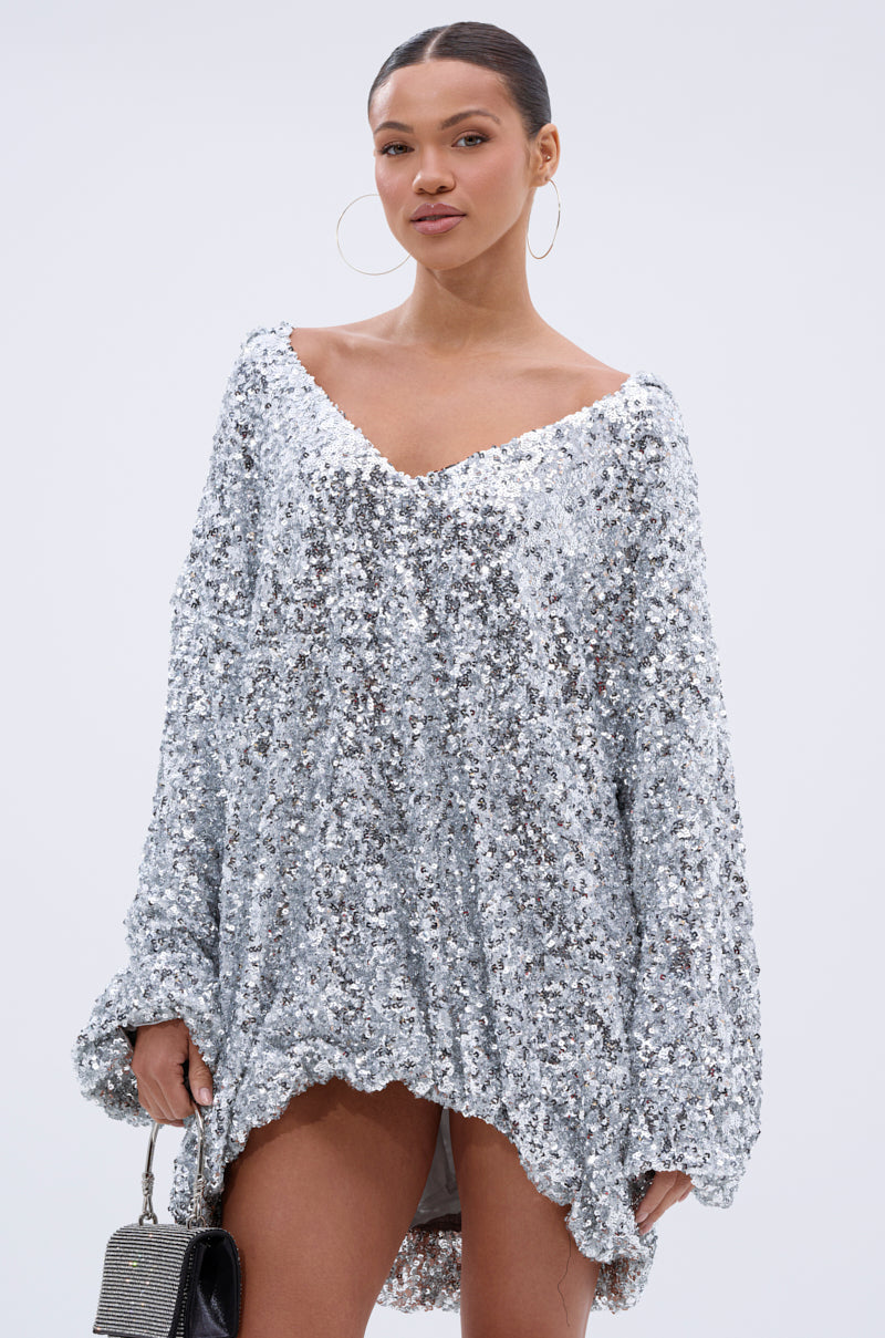 A woman wears the GISELLE SEQUIN SWEATSHIRT DRESS IN SILVER with large hoop earrings and a small textured handbag, standing confidently against a plain light background.