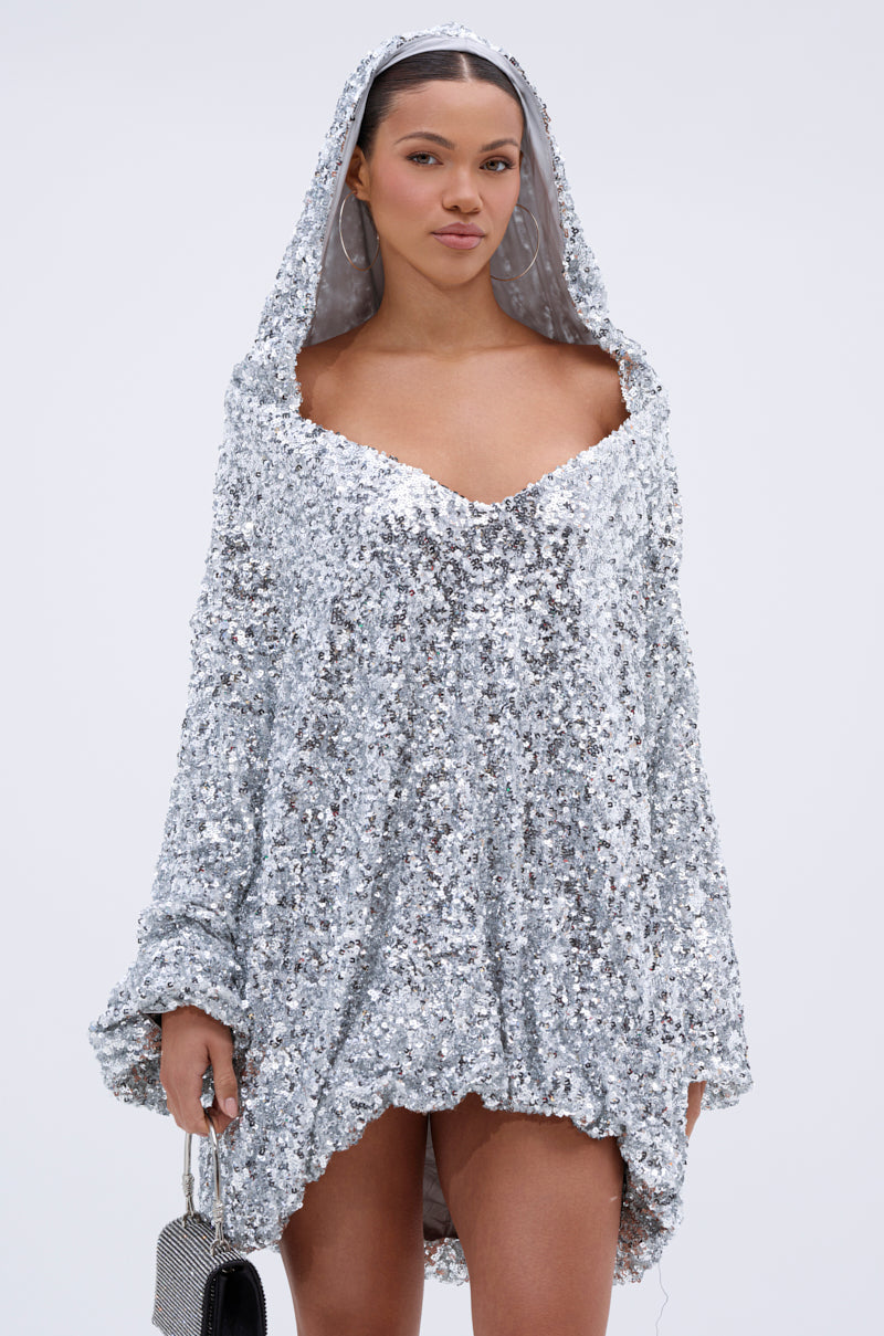 A woman stands confidently against a plain background, wearing the GISELLE SEQUIN SWEATSHIRT DRESS IN SILVER with a deep neckline and small silver handbag—a glamorous choice for parties.